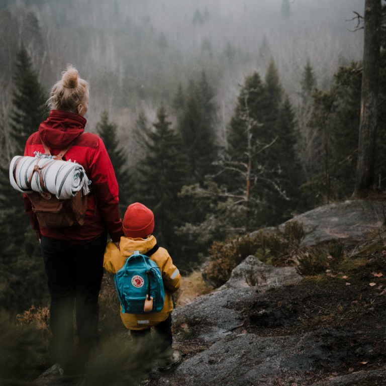 Äiti ja lapsi käsi kädessä kalliolla metsässä utuisessa syysmaisemassa.