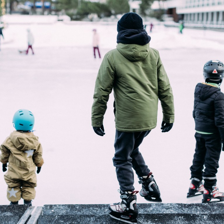 Kolme lasta starttaamassa luistelukentän reunalta luistelemaan.