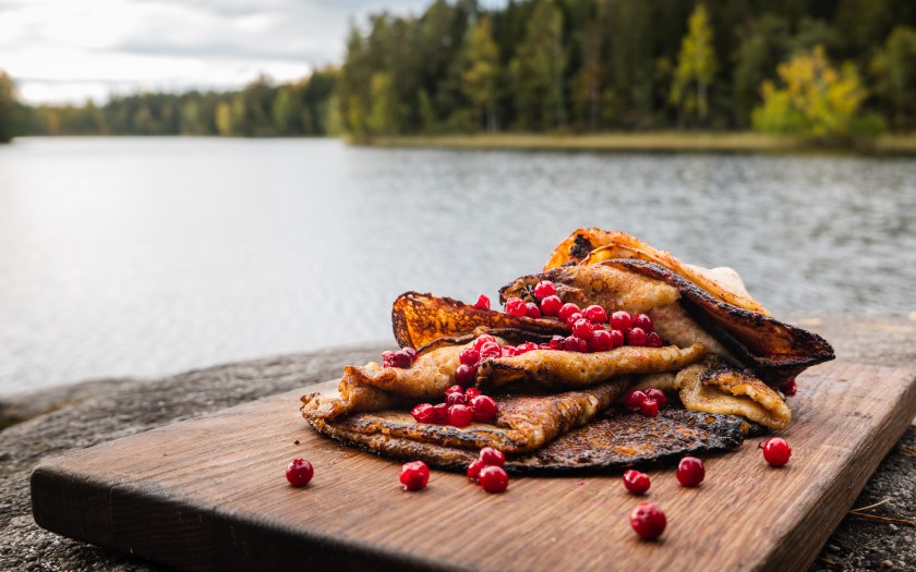 Juuri paistettuja lettuja karpaloilla ja kinuskikastikkeella puulankun päällä järven rannassa.