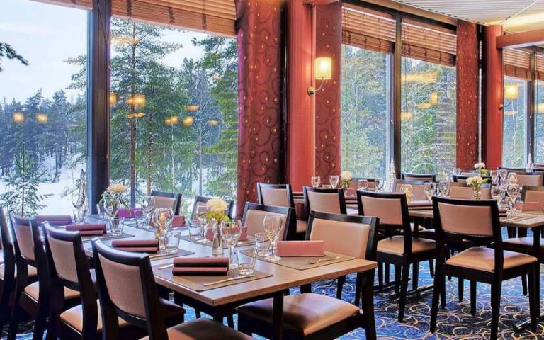 A restaurant hall with groups of tables and whose large windows overlook the snowy forest.